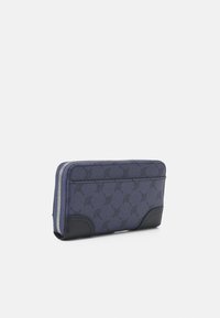 Portefeuille texturé bleu avec un motif logo, doté d'une base en cuir noir et d'une fermeture éclair argentée. Forme rectangulaire avec un design élégant.