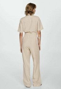Play suit beige con maniche corte, vita aderente e gambe larghe. Realizzato in tessuto leggero, presenta un design semplice senza motivi o dettagli.