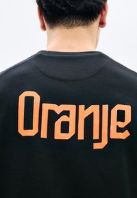 Άτομο με κοντά κατσαρά μαλλιά που φοράει μαύρο πουκάμισο με μεγάλα έντονα πορτοκαλί γράμματα «Oranje» στην πλάτη.