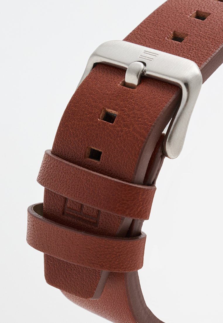 Correa de reloj de cuero marrón con una superficie texturizada y hebilla de metal plateado. Presenta dos fijaciones ajustables y agujeros cuadrados para el ajuste.