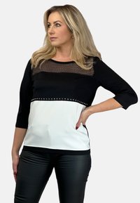 Czarna i biała bluzka z siateczkowym dekoltem, rękawami o długości 3/4 oraz ćwiekowanym akcentem w talii. Wykonana z gładkiego materiału o wysmuklonym kroju.
