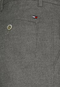 Grau gemischte Wollhose mit glatter Textur, ausgestattet mit einer vorderen Tasche und einem Knopfverschluss. Ein kleines Logo in Rot, Weiß und Blau ist sichtbar.