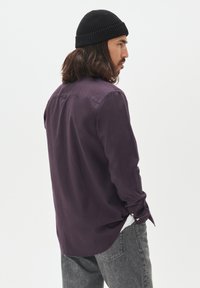 Man med långt hår som bär en svart mössa, mörklila skjorta och grå jeans, vänd åt sidan mot en enkel ljus bakgrund.