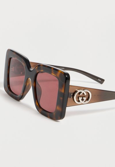 Τετράγωνα γυαλιά ηλίου με σχέδιο χελώνας, μοβ φακούς και χρυσό λογότυπο Gucci πάνω σε καφέ διάφανα πλαίσια.