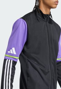 Schwarze Sportjacke mit lila Ärmeln. Verfügt über einen hohen Kragen, einen Reißverschluss vorne, ein weißes Logo und grüne Akzentstreifen an den Ärmeln.