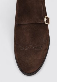 Zapato de ante marrón con punta redondeada, costura en forma de ondas y correa lateral con hebilla dorada. Suela plana de goma negra.