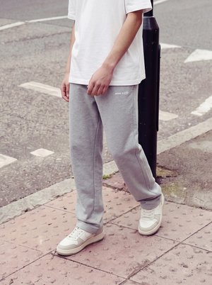 Person iført hvide sneakers, grå joggingbukser og en hvid t-shirt står på en tekstureret fortov nær en sort pæl.