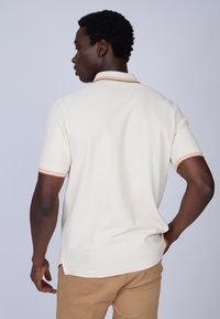 Polo shirt crème à manches courtes avec des rayures marron sur le col et les manches. Il dispose d'une coupe décontractée et d fentes sur les côtés pour plus de confort.