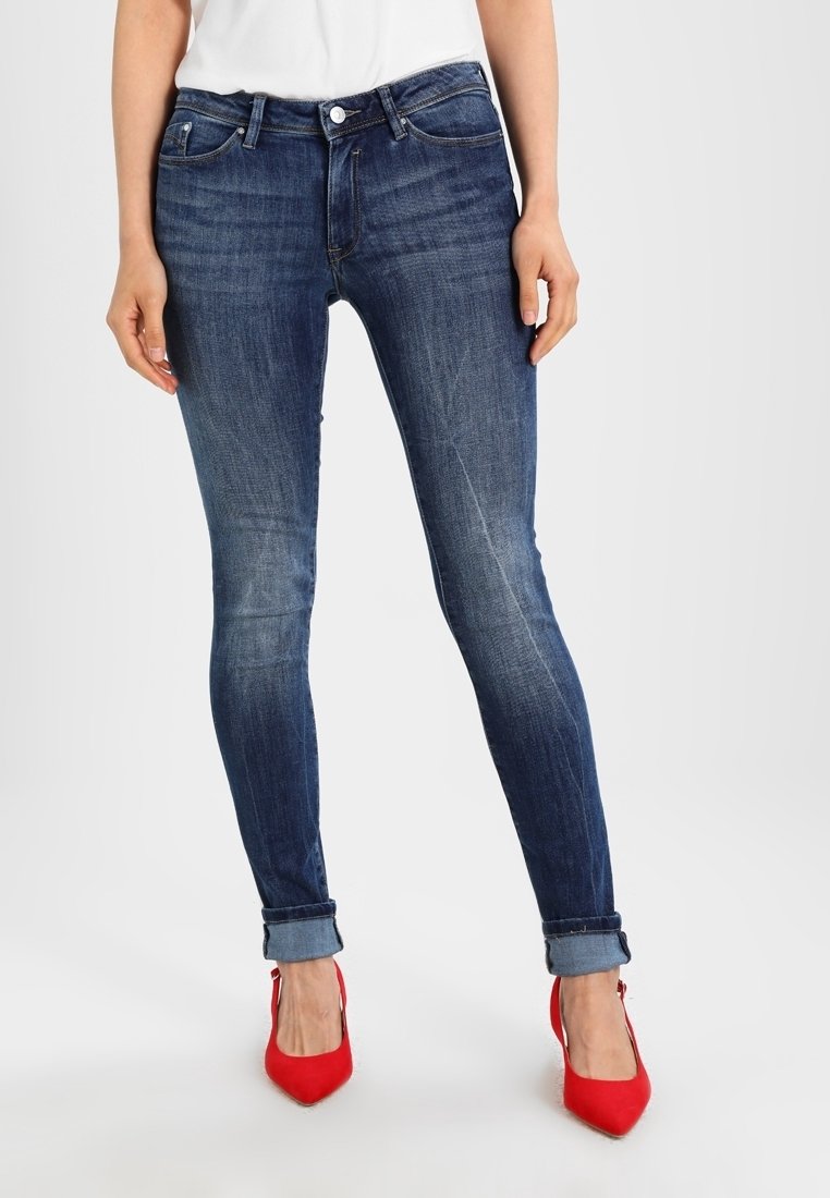 Vaqueros ajustados de denim azul oscuro, con una cintura de altura media, bolsillos frontales y un ligero desvanecimiento. Los puños doblados revelan unos tacones rojos.