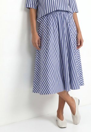 Žena v modro-bielej sukni s vertikálnymi pruhmi a zodpovedajúcom topom, stojaca s bielymi slip-on topánkami pri bielej stene.