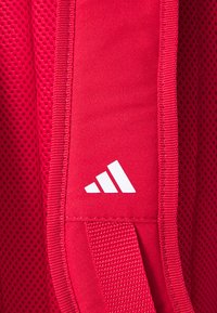 Red backpack featuring textured mesh and fabric materials, with stitched accents and a white Adidas logo on the side.
