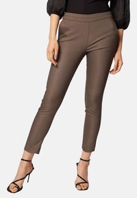 Pantalons marron sur mesure avec une coupe slim, devant plat et longueur cheville courte, associés à des talons hauts noirs à lanières. Texture lisse et détails minimaux.