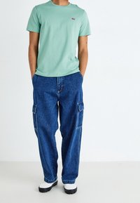 T-shirt en coton vert menthe avec un petit logo rouge sur la poitrine, associé à un pantalon cargo en denim bleu ample avec des poches latérales.