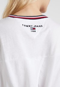 Bílý bavlněný tričko s červeným a modrým pruhovaným žebrovaným výstřihem a vyšívaným logem "TOMMY JEANS 1985" na zadní straně, s vý�řezem ve tvaru V.
