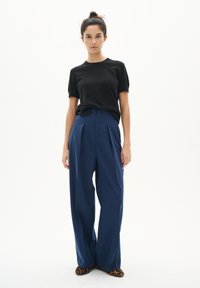 Schwarzes T-Shirt mit kurzen Ärmeln, kombiniert mit hochgeschnittenen, lockeren blauen Hosen mit Falten und Leopardenmuster-Schuhen.