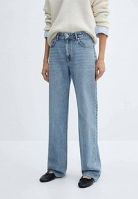 Jeans de tiro alto de pernera recta en azul claro combinados con bailarinas negras y un suéter crema con puños de camisa azul visibles.