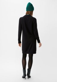 Schwarzes Strickkleid mit langen Ärmeln, gerippten Bündchen und Saum. Getragen mit schwarzen Strumpfhosen und Stiefeletten. Eine grüne Strickmütze sorgt für Kontrast.