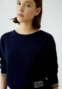 Dunkelblaues Strickoberteil mit Rundhalsausschnitt und dreiviertellangen Ärmeln. Mit gestreiften Bündchen und einem rechteckigen Patch mit "OUI" in Weiß.