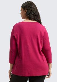 Frau mit langem, dunklem Haar, die ein leuchtend pinkfarbenes Langarmoberteil und schwarze Hosen trägt, mit dem Rücken zu einem schlichten hellen Hintergrund gewandt.