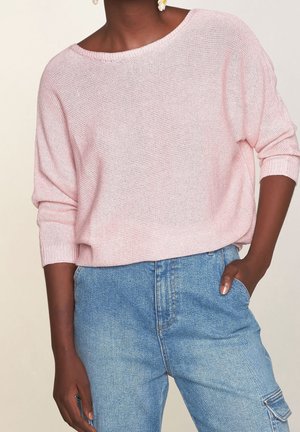 Tummaihoinen henkilö, joka pukeutunut vaaleanpunaiseen löyhästi neulottuun neulepuseroon ja sinisiin korkeavyötäröisiin farkkuihin, toinen käsi taskussa.