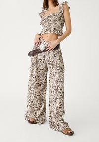 Vrouw in een bijpassende paisley-print crop top en wijde broek met een gevlochten bruine riem en platform sandalen.