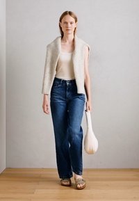 Modella che indossa un gilet beige senza maniche in tessuto sopra una canottiera bianca, abbinato a jeans a gamba larga blu e sandali marrone chiaro, mentre tiene una borsa bianca.