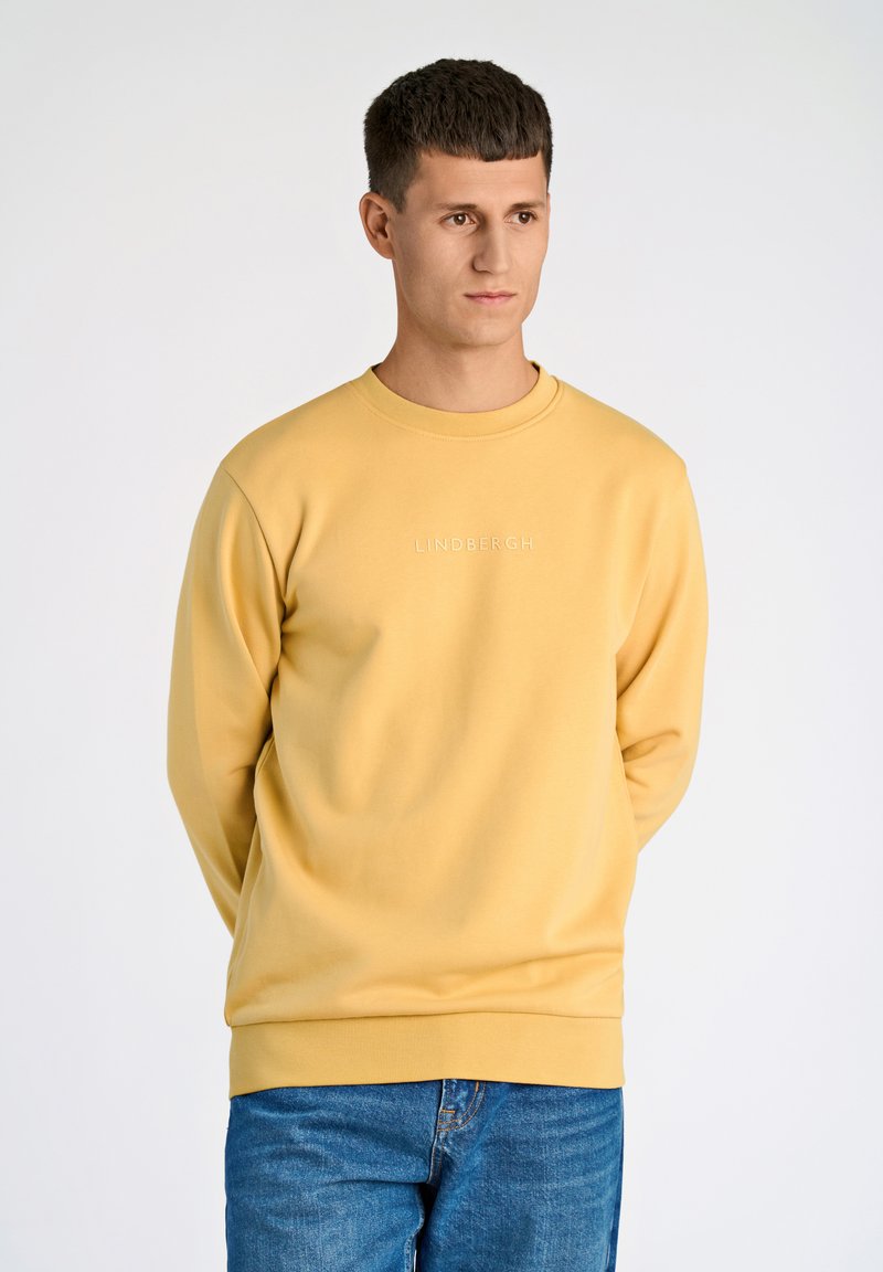 Felpa gialla con scollo rotondo, maniche lunghe e logo ricamato "LINDBERGH" sul davanti. Indossata con jeans blu.