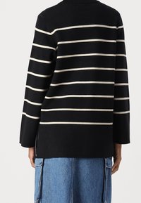 Personne portant un haut noir à manches longues avec des rayures horizontales blanches, associé à un pantalon cargo en denim bleu, vue de dos.