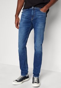 Blå denimjeans med en smal passform, som har fem fickor, kontrasterande sömmar och en knappstängning. Bärs med svarta kanvas sneakers.