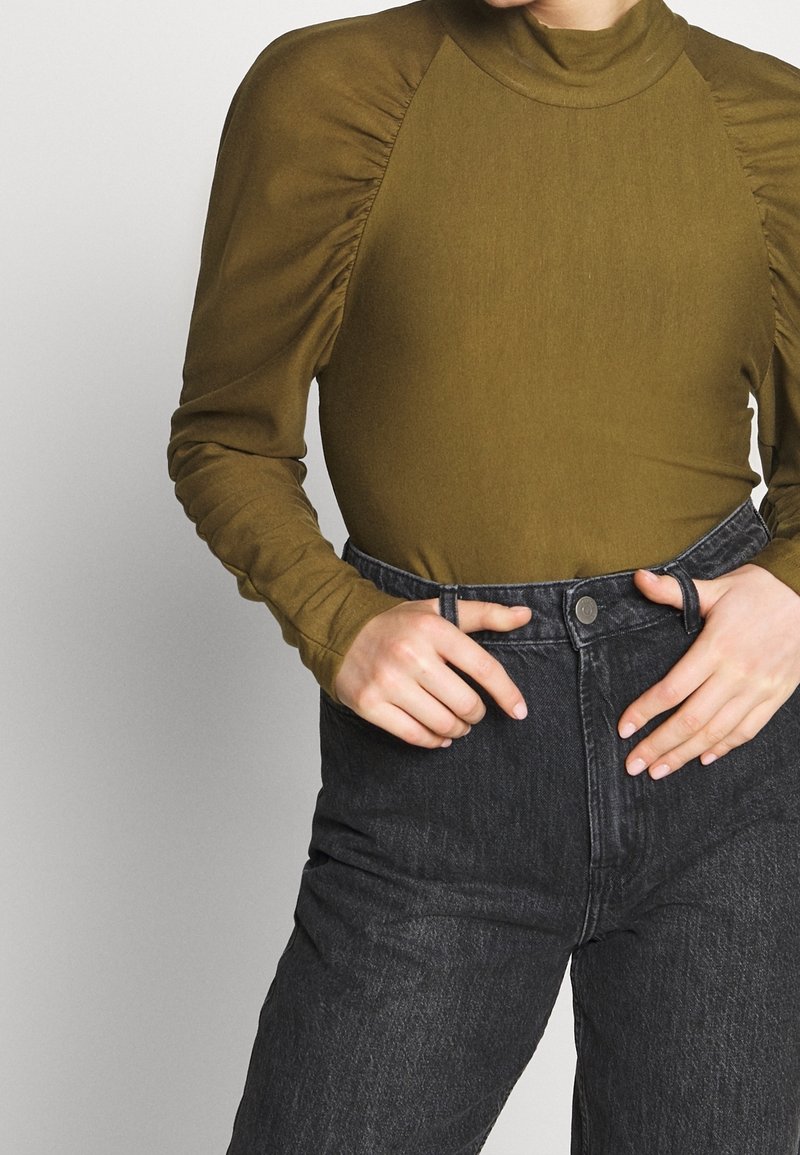 Personne portant un haut à manches longues vert olive avec des épaules froncées et un jean noir taille haute, les mains ajustant la taille.