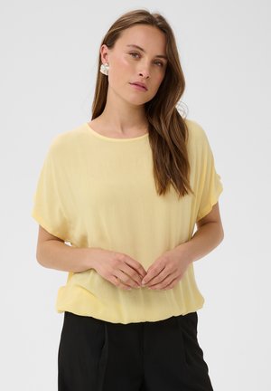Donna con lunghi capelli castani che indossa una camicetta giallo chiaro a maniche corte e pantaloni neri, in piedi con le mani delicatamente intrecciate.