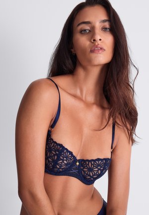Femme aux longs cheveux bruns portant un soutien-gorge en dentelle bleu marine avec des motifs floraux sur un fond gris clair uni.