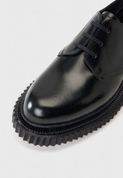 Μαύρο γυαλιστερό δερμάτινο παπούτσι με χοντρή, ραβδωτή σόλα και μαύρα κορδόνια, κοντινή λήψη της μύτης και του επάνω μέρους σε φωτεινό φόντο.