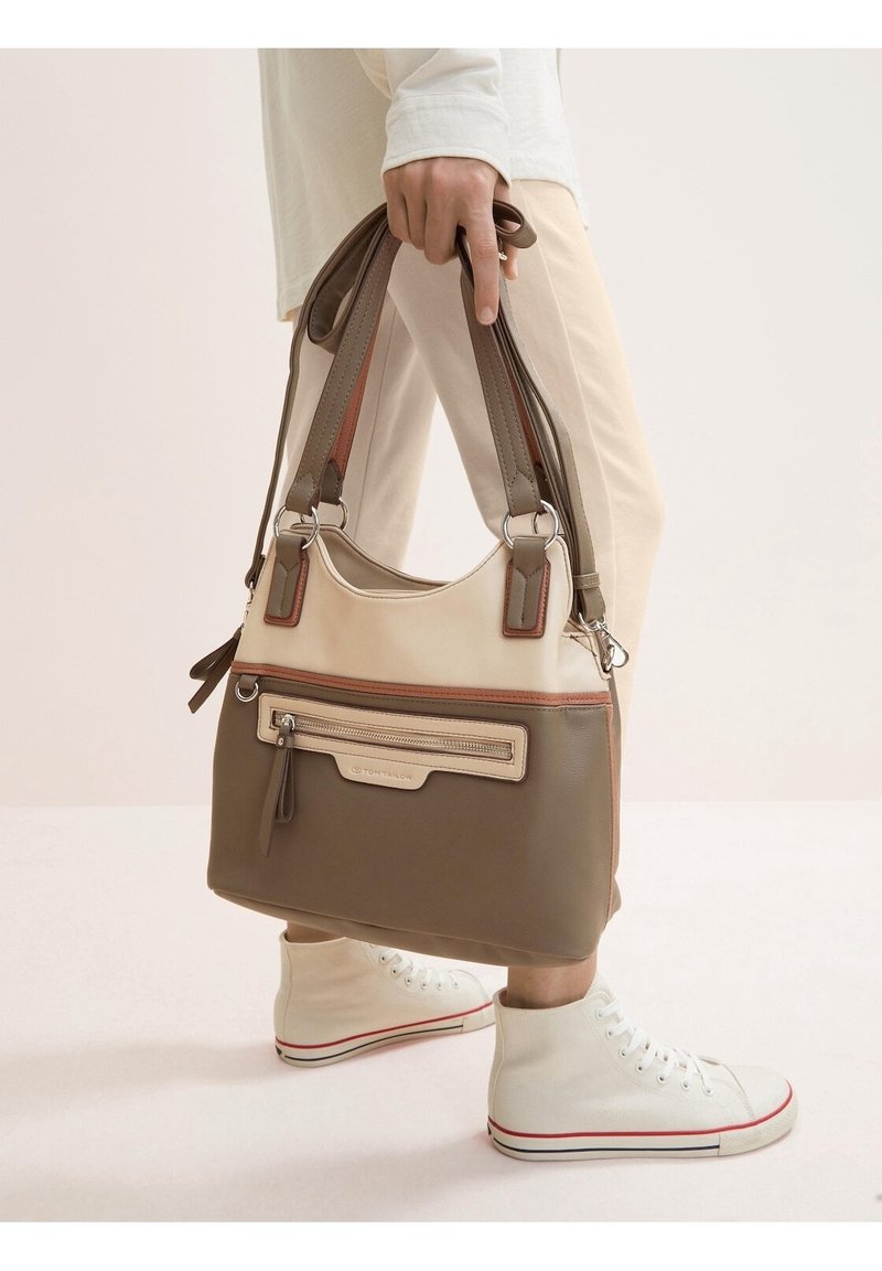 Personne portant un pantalon blanc et des baskets montantes blanches tenant un sac à main bicolore beige et marron par ses anses.
