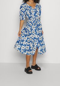 Modré květinové šaty s bílými a černými květy, výstřih do V, krátké puffové rukávy a zavazování v pase, doplněné černými platformovými sandály.