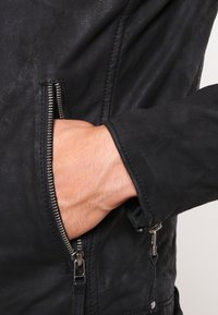 Hand med ljus hudton inskjuten i den öppna fickan på en svart läderjacka med synlig dragkedja och stygn.