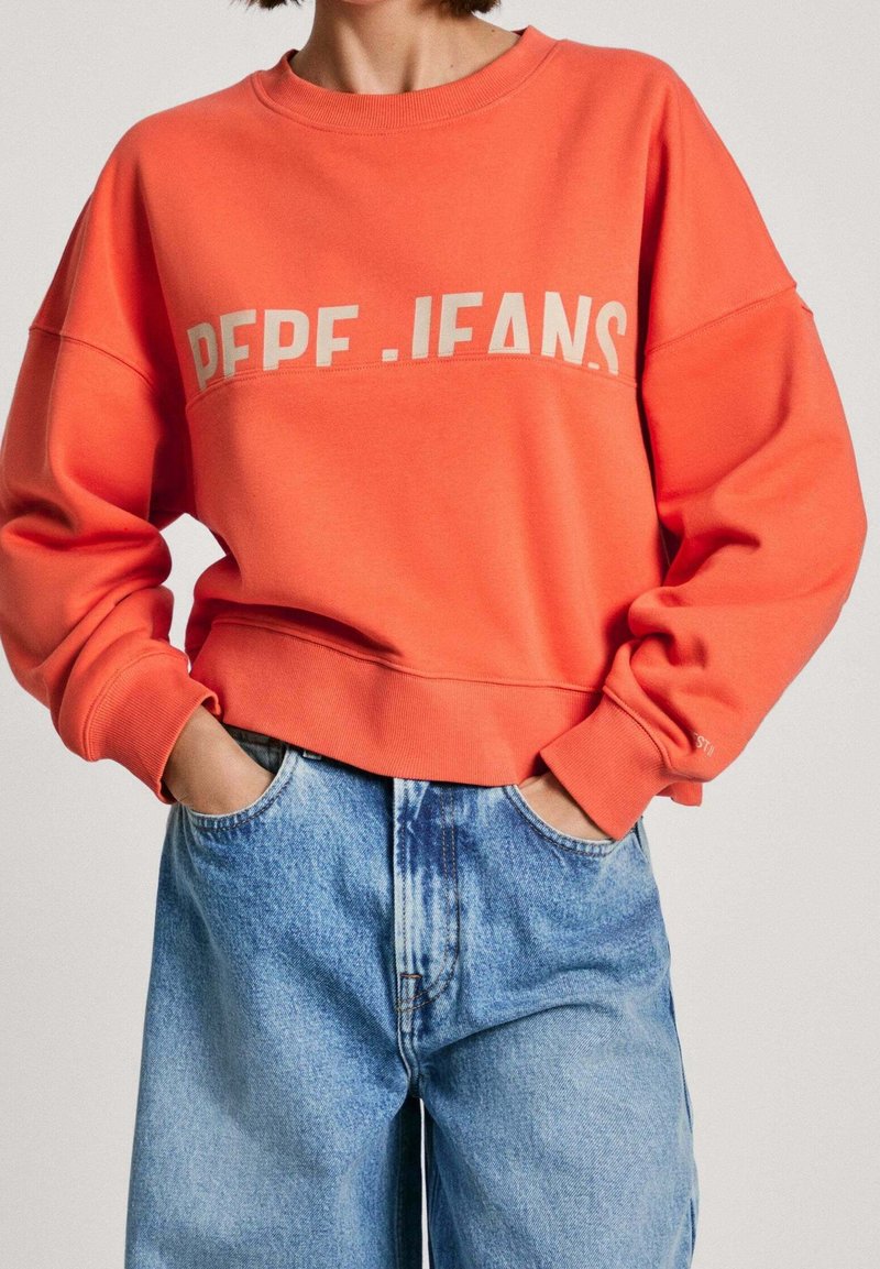 Oranžová mikina s kulatým výstřihem a žebrovanými manžetami, na hrudi s velkým bílým logem "PEPE. JEANS". Nosená s světle modrými džíny.