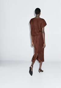 Hnědé semišové šaty s volným a oversized střihem, krátké rukávy, délka po kolena a boční rozparek. Vytvořeno v kombinaci s černými lodičkami.