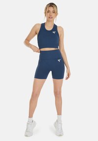 Marineblaue Sport-BH- und Shorts-Ausstattung, gerippte Textur, Logodetails auf beiden Teilen, hoher Taillenbund bei den Shorts, kombiniert mit weißen Turnschuhen.