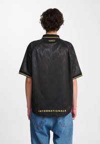 Μαύρη φανέλα με υφασμάτινο σχέδιο, κίτρινες λεπτομέρειες στον γιακά και τα μανίκια, και την ένδειξη "INTERNATIONALS" τυπωμένη στο στρίφωμα.