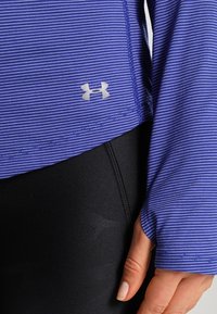 Lila långärmad träningströja med smala vita ränder, med en silverlogga vid fållen. Kombinerad med svarta tights.