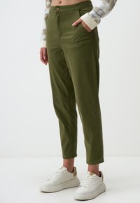 Pantaloni verde oliva, a gamba affusolata con vita alta, tasche laterali e chiusura con bottone, abbinati a sneakers bianche e a un maglione a righe texture.