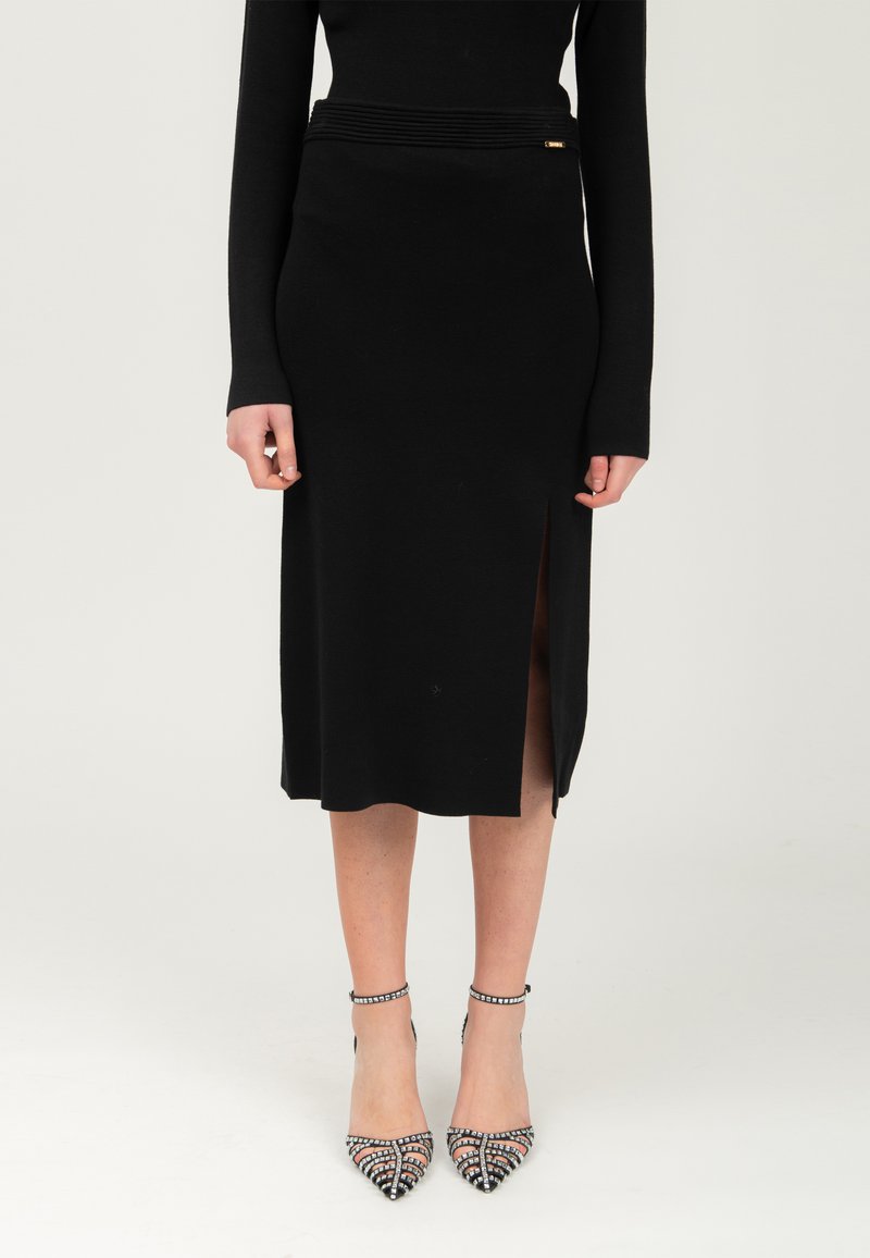 Jupe en tricot noir avec une fente sur le côté et une taille texturée. Portée avec des talons aiguilles noirs ornés, avec des brides autour de la cheville.