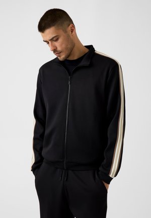 Uomo che indossa una giacca sportiva nera con zip, con strisce beige e bianche sulle maniche, mani nelle tasche, guarda in basso, sfondo semplice.