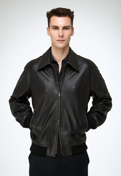 Joven con una chaqueta negra de cuero con cremallera y las manos en los bolsillos, de pie contra un fondo blanco liso.