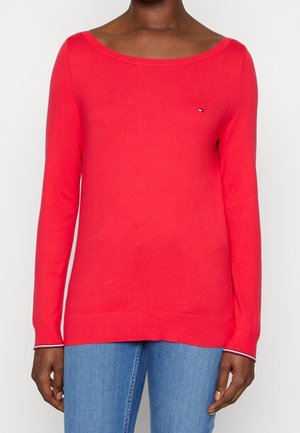 Kobieta w jasnoczerwonym swetrze z długim rękawem i dekoltem typu łódka oraz małym logo, w połączeniu z niebieskimi dżinsami.