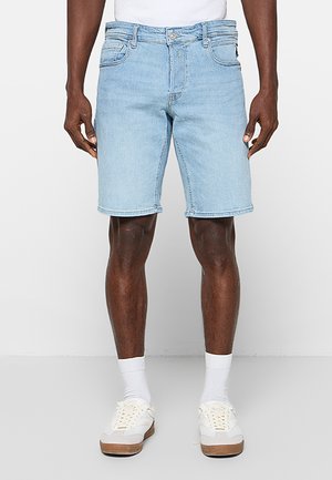 Mężczyzna ubrany w jasnoniebieskie jeansowe szorty, białe skarpetki i białe trampki, stojący na jednolitym jasnym tle.