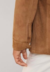 Chaqueta de ante perforada marrón con puños doblados y costuras visibles. El material presenta una textura suave y cuenta con un diseño casual.