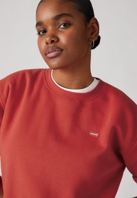 Jeune femme à la peau foncée et aux cheveux en chignon, portant des boucles d'oreilles en or et un sweat-shirt rouge Levi's sur une chemise blanche.