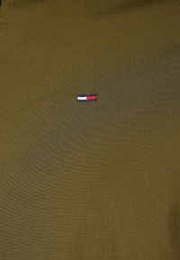 Olivgrön skjorta gjord av mjukt tyg, med en liten broderad röd, vit och blå logotyp placerad på vänster sida.